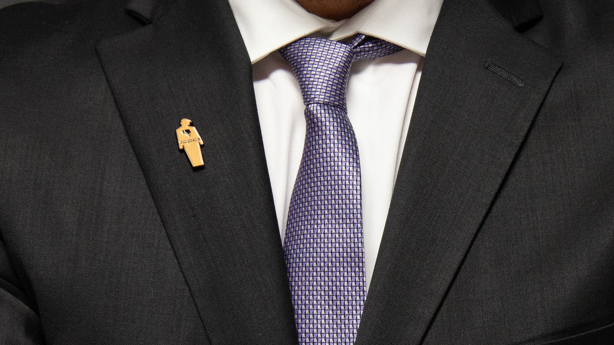 Closeup of a 1.5" tall tiny trump enamel pin on a men's suit lapel. tiny trump has a thumbs down sign in the middle of its chest