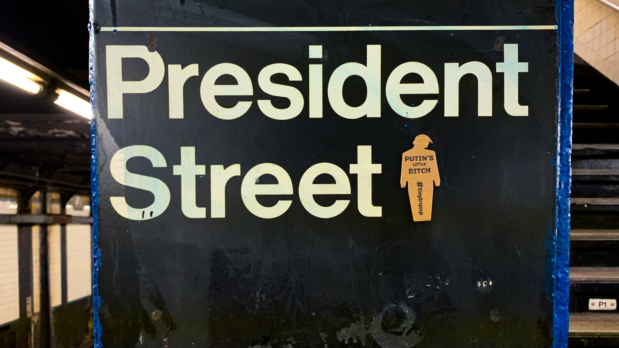 tiny trump with the slogan "Putin's Little Bitch" stuck to a NYC subway sign indicating "President Street"