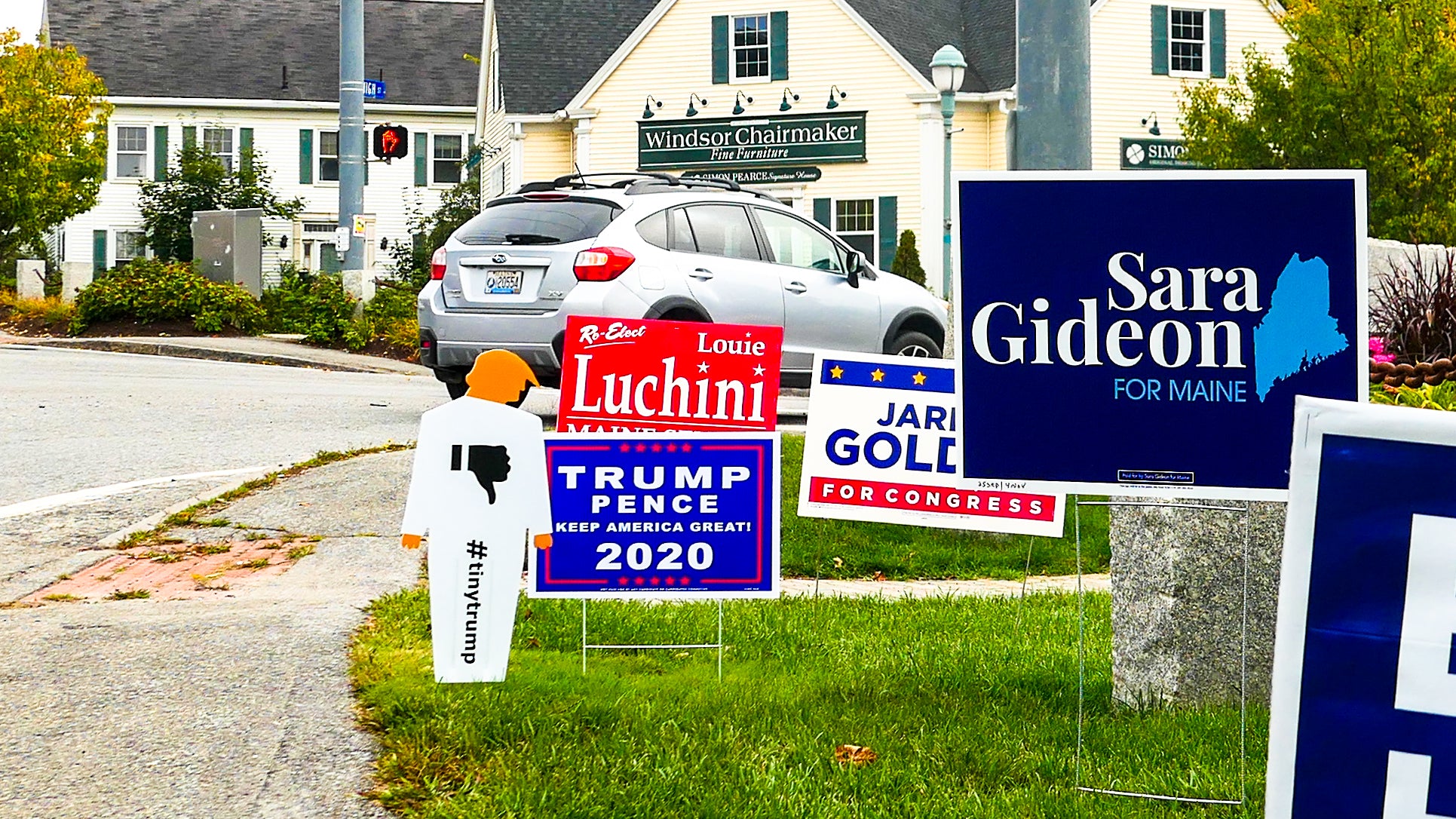 two foot tall tiny trump lawn sign (white coroplast trump cutout with orange head and hands) with a thumbs down sign in the chest standing amidst four other traditional political lawn signs on a street corner