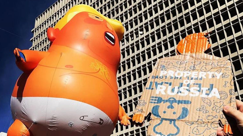 A two foot tall, cardboard tiny trump at a protest held up high next to the trump baby blimp