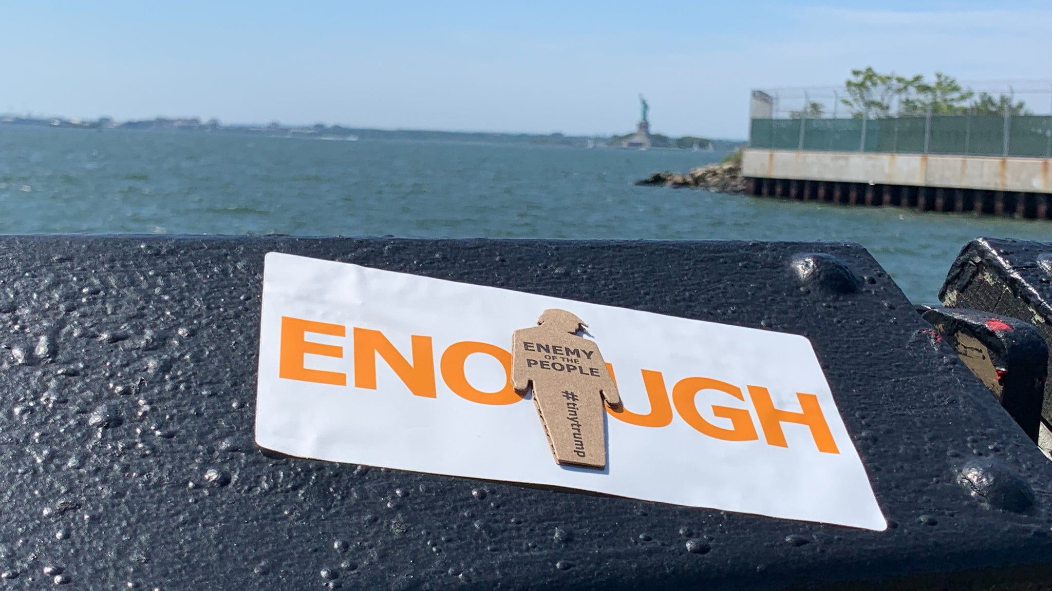 "ENOUGH" tiny trump sticker measuring 3.75" x 7.5" shown stuck to a railing with the Statue of Liberty in the background