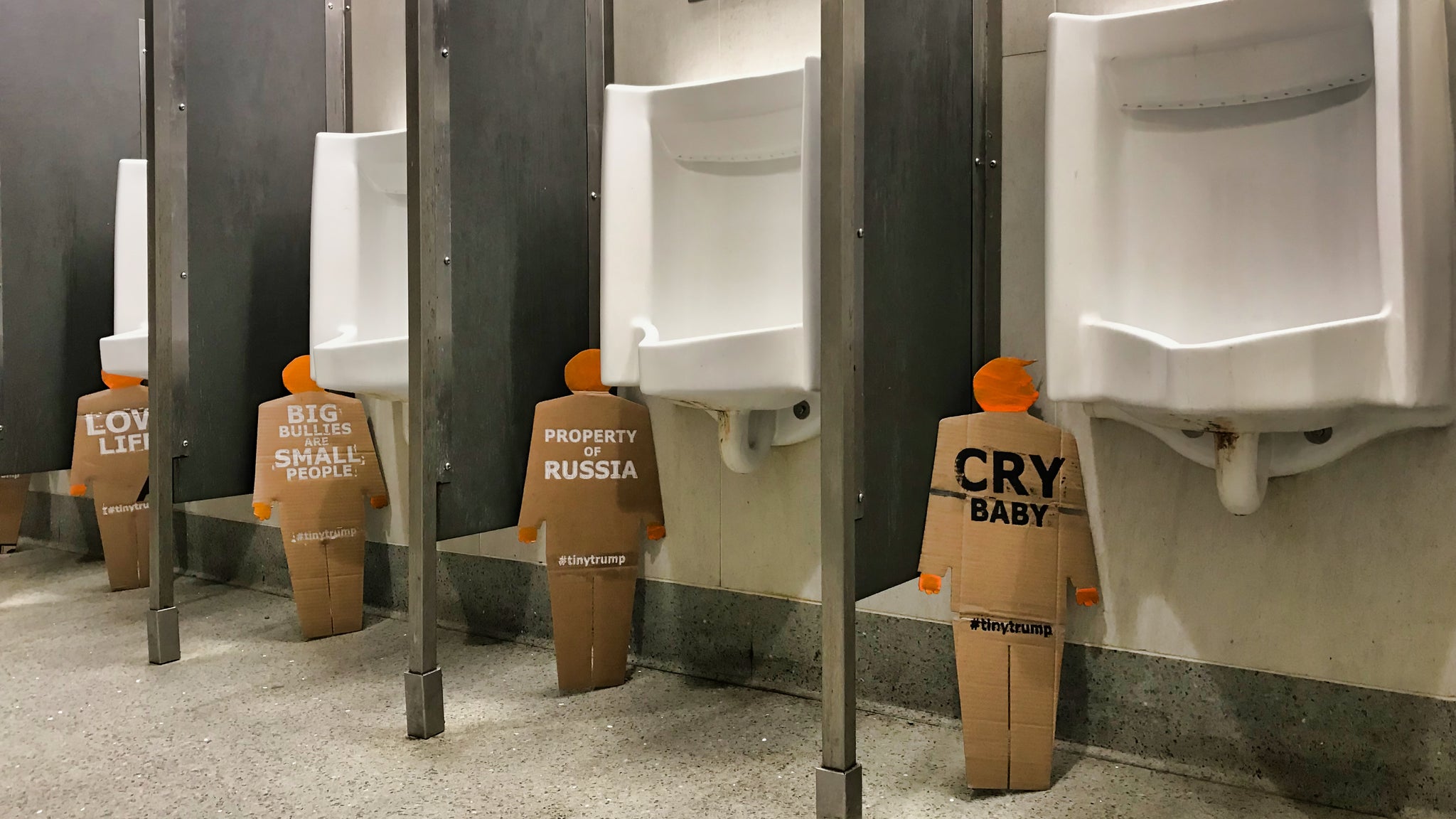 Four, two foot tall, cardboard tiny trumps, each standing next to a urinal in a row of urinals