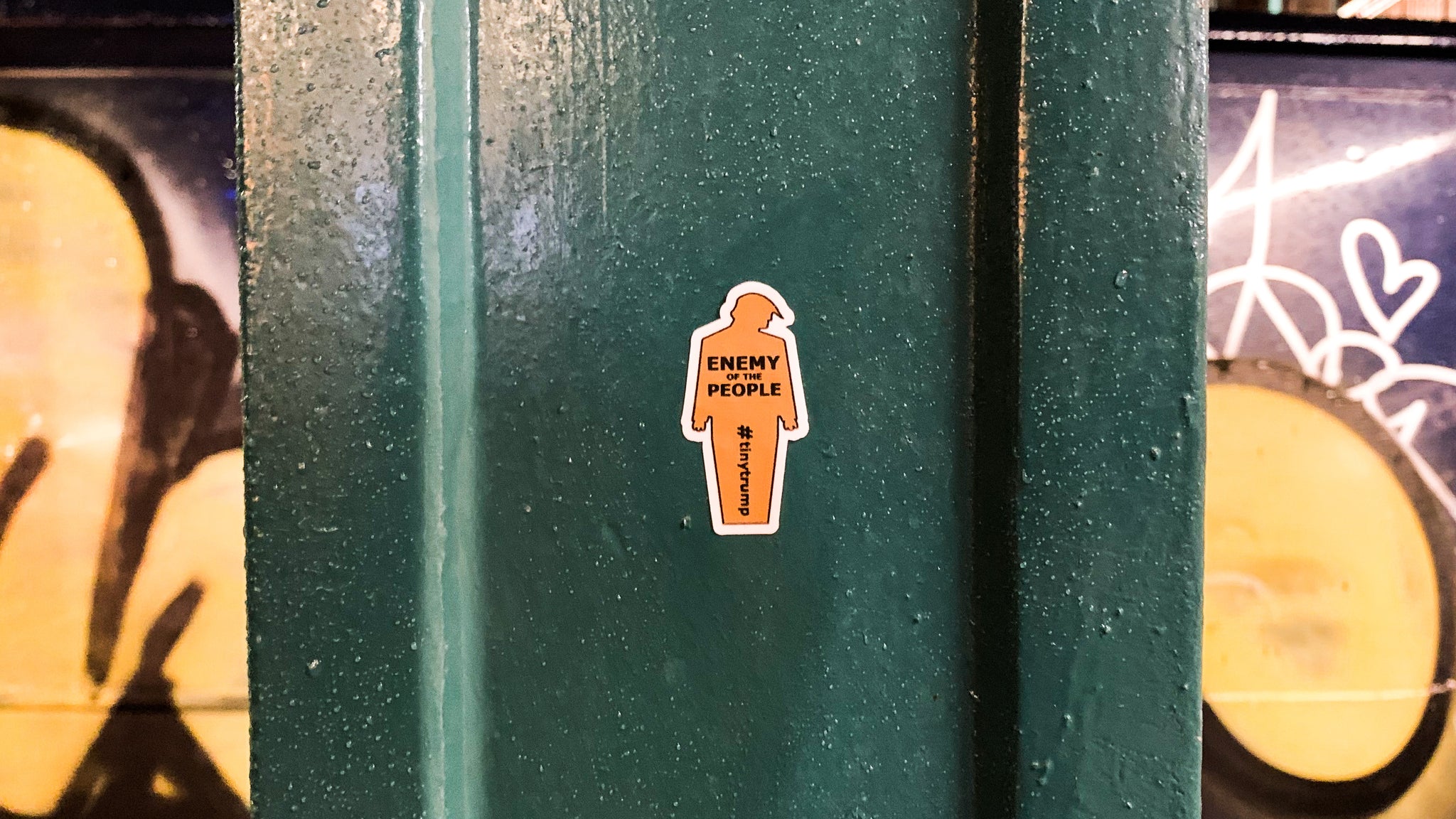 A two inch tall tiny trump sticker with the slogan Enemy of the People stuck on a green column