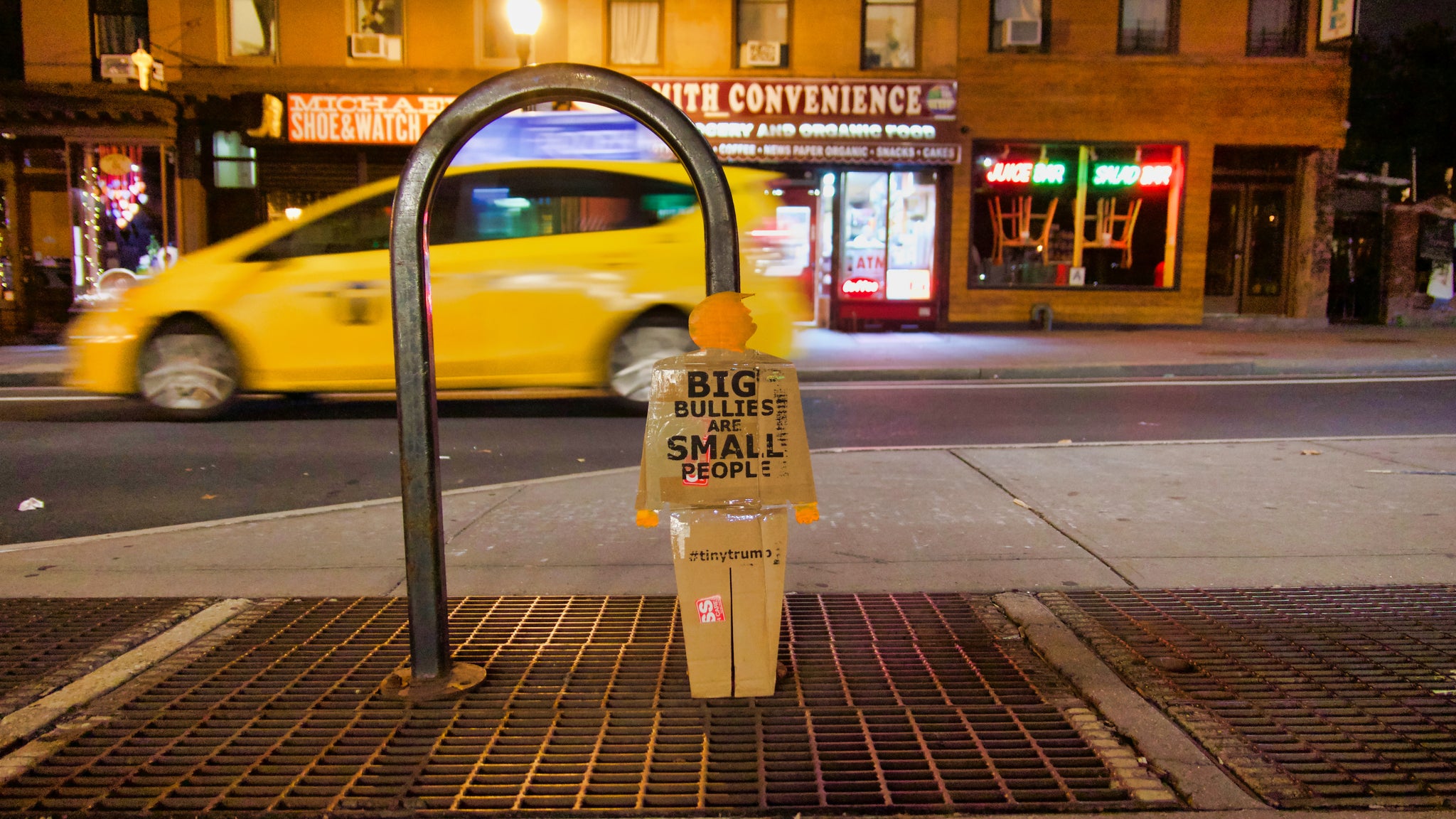 A two foot tall, cardboard tiny trump with the slogan "Big Bullies Are Small People", leaning against a bike rack on the street in New York City