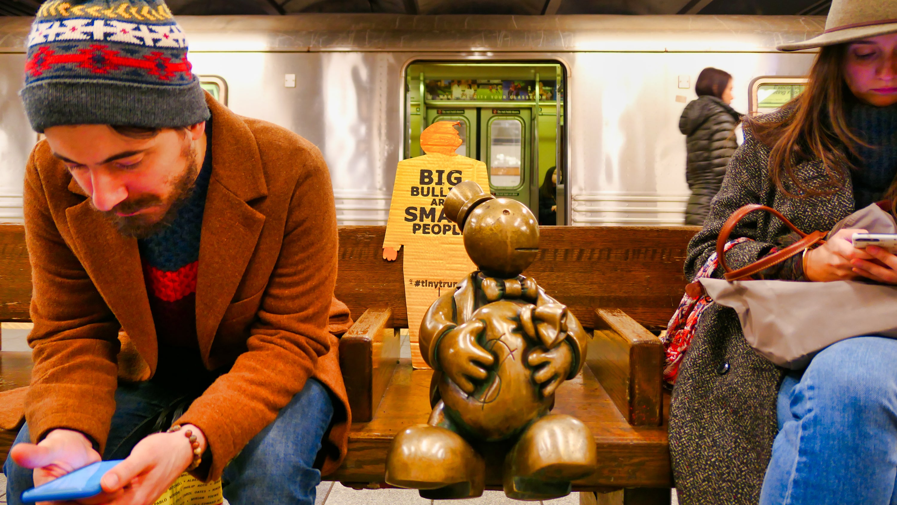 A two foot tall tiny trump sitting on a bench in the New York City subway, between two people reading