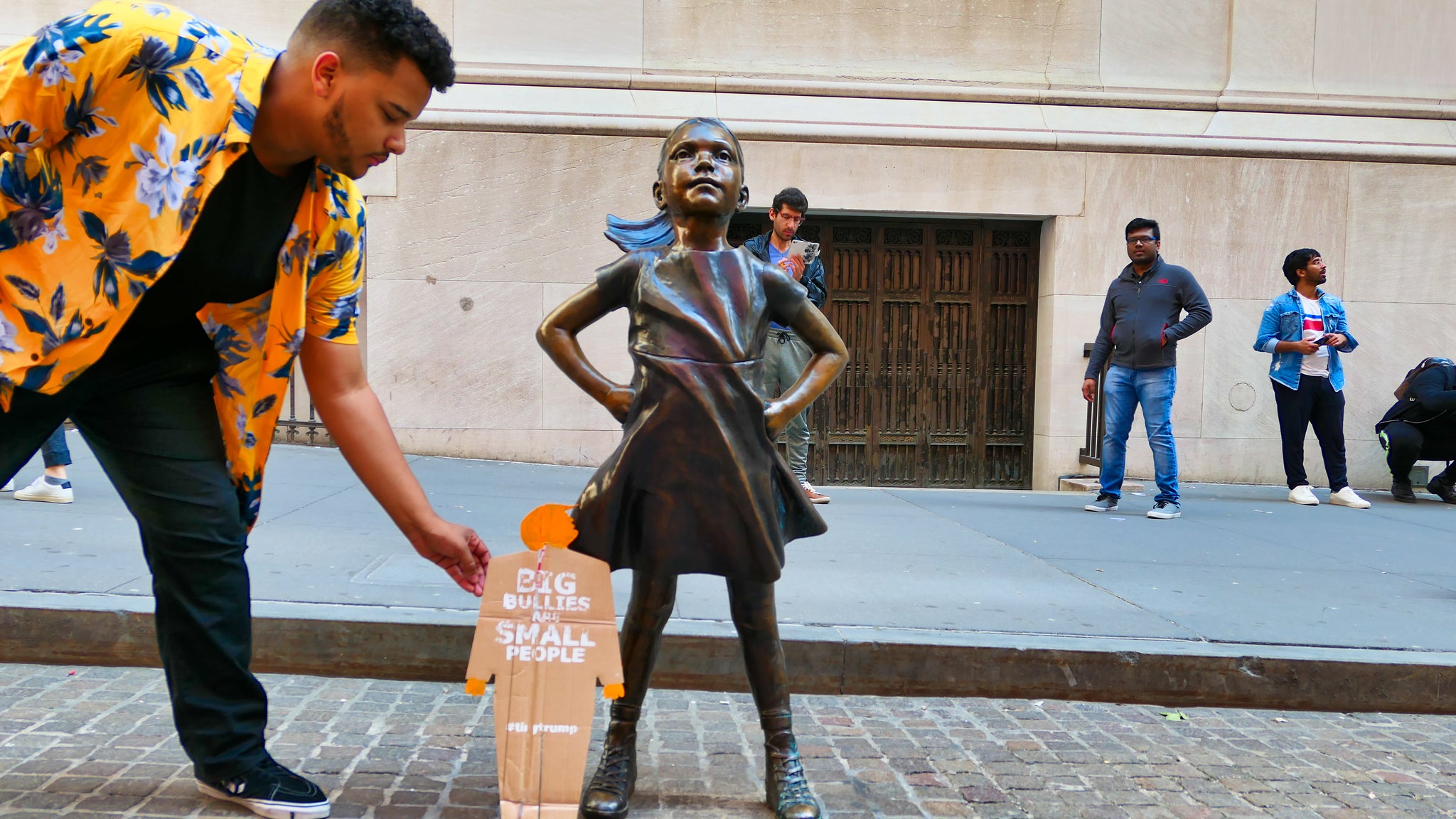 A two foot tall, cardboard tiny trump with the slogan "Big Bullies Are Small People" looking up a the Fearless Girl statue in lower Manhattan