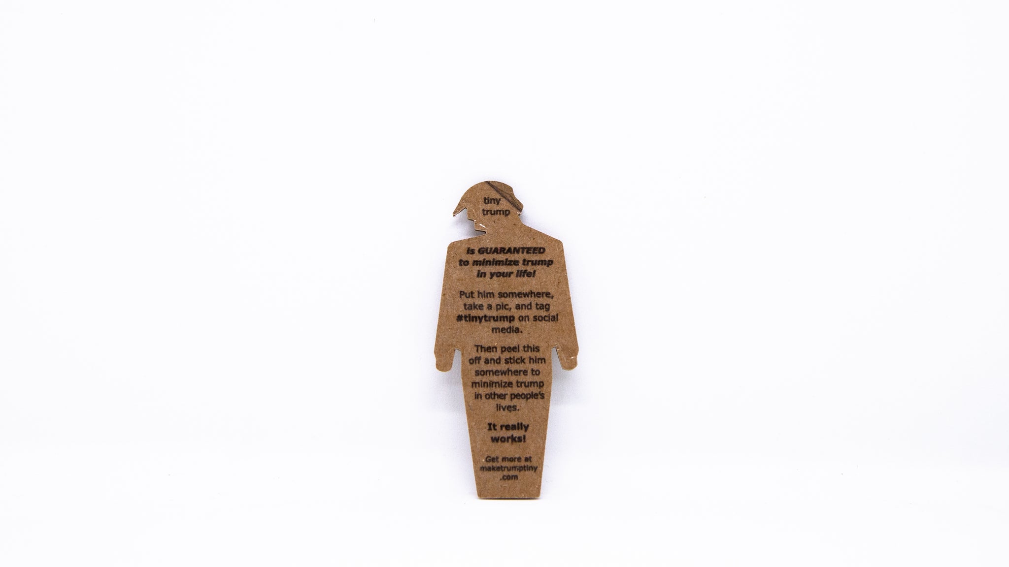 The back of a tiny trump showing a peel off label with the following text: "tiny trump is guaranteed to minimize trump in your life! Put him somewhere, take a pic, and tag #tinytrump on social media. Then peel this off and stick him somewhere to minimize trump in other people's lives. It really works! Get more at maketrumptiny.com"